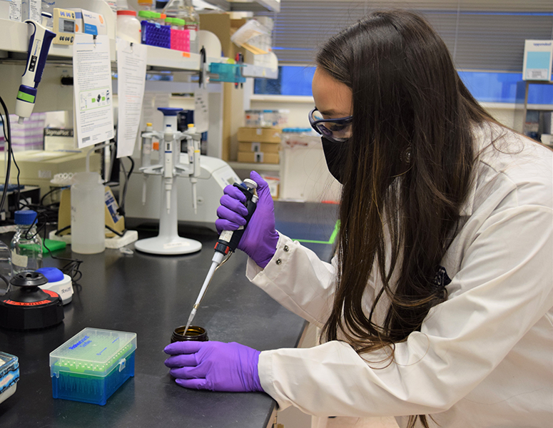 Samantha Wade stands in a research lab wearing a white coat with purple gloves holding a pipette